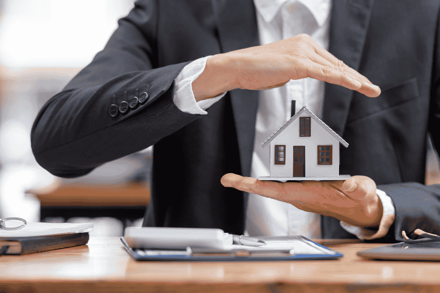 Protection du patrimoine : homme d’affaires protégeant une maison miniature symbolisant la sécurité des biens et des proches.