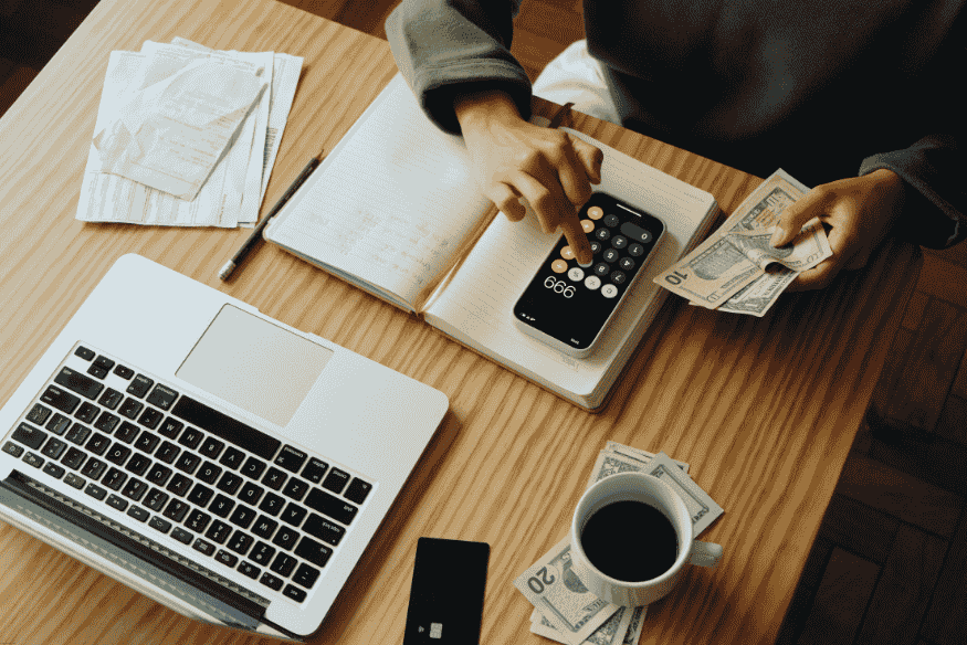 Personne utilisant une calculatrice sur smartphone pour gérer de l’argent liquide, avec ordinateur et carnet de notes.