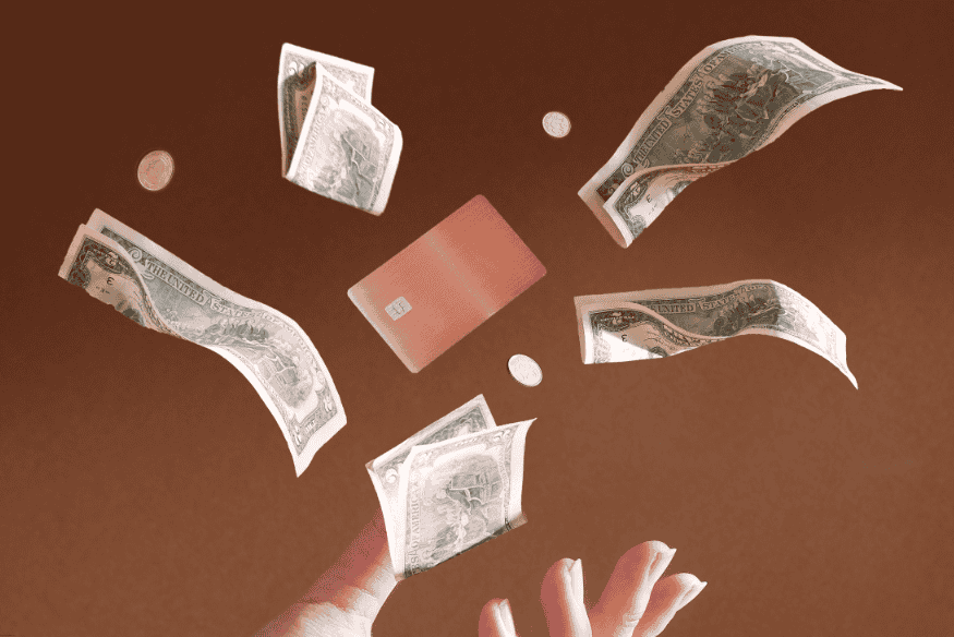 Main lançant des billets de banque et une carte bancaire en l’air sur fond marron.
