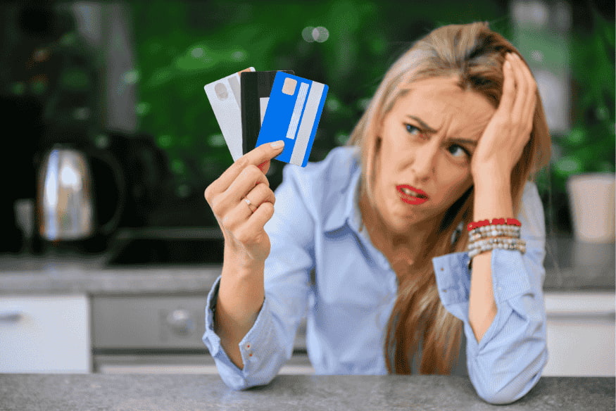 Femme stressée tenant plusieurs cartes bancaires dans une cuisine, symbole d’un refus de paiement ou surcharge de dettes.