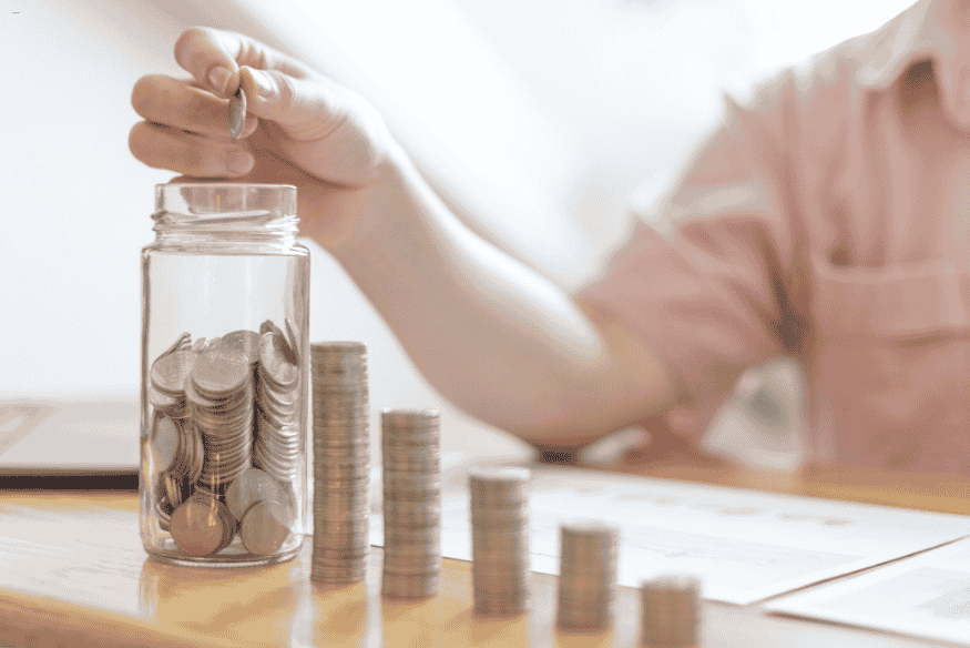 Main plaçant une pièce dans un bocal rempli de pièces, avec piles de monnaie alignées sur une table.