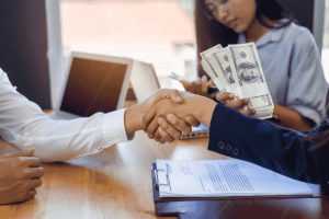 Poignée de main entre deux personnes après un accord de prêt, avec une femme tenant des billets de banque à l’arrière-plan.