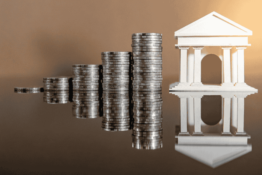 Empilement de pièces de monnaie devant un bâtiment bancaire, illustrant les bénéfices réalisés par les banques grâce aux découverts.