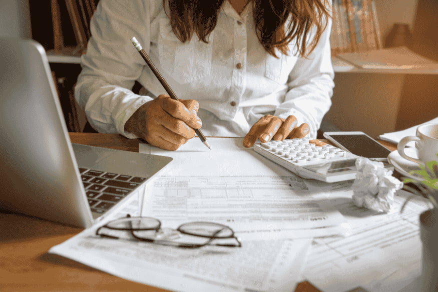 Femme calculant son budget avec des documents financiers, un ordinateur portable et une calculatrice sur un bureau.