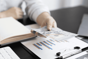 Personne tendant des billets de banque au-dessus d’un graphique financier sur une table.