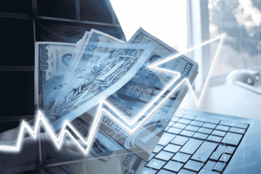 Billets de banque en main avec courbe de croissance devant un ordinateur, illustrant les décisions financières en 2025.