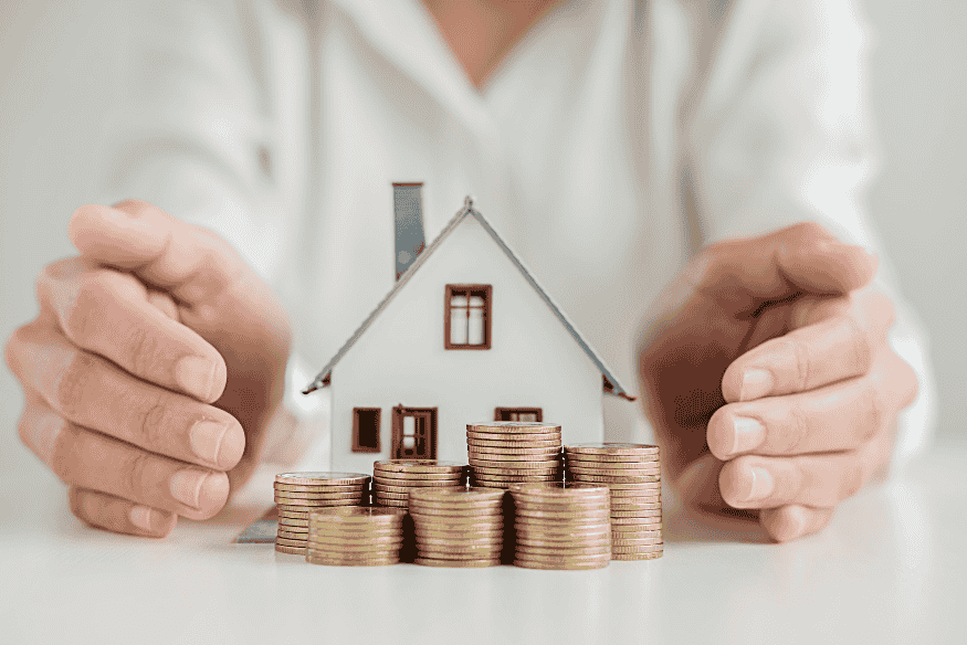 Modèle de maison entouré de pièces empilées et de mains protectrices, représentant l’épargne logement avec un PEL ou CEL en 2025.