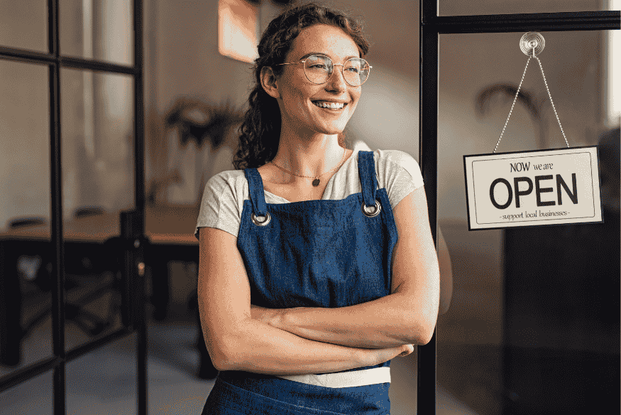 Jeune auto-entrepreneuse souriante devant son commerce ouvert, symbole de réussite grâce à un prêt d’honneur.