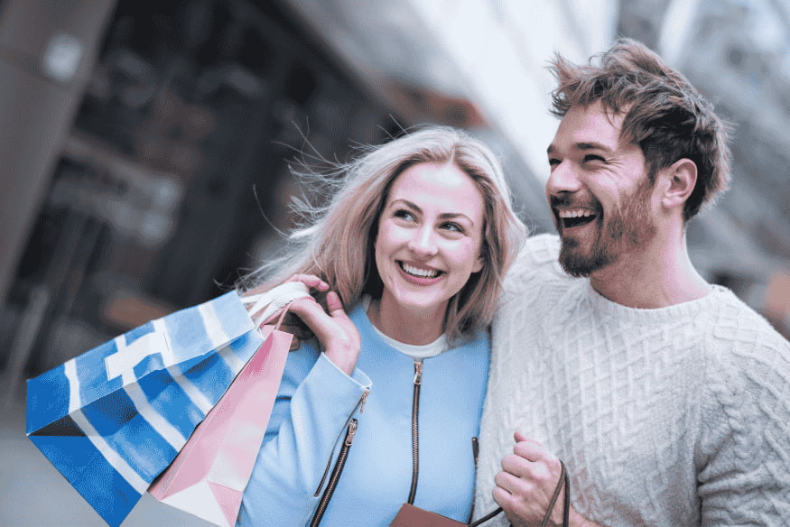 Couple souriant faisant du shopping avec plusieurs sacs, illustrant une dépense motivée par l’euphorie.